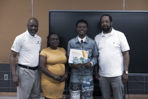 Four people pose indoors; one young man in the center holds a certificate and smiles at the camera.