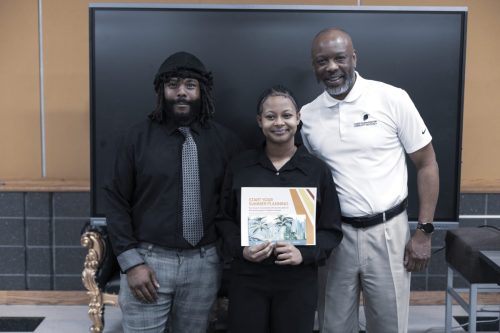 Three people pose indoors; the woman in the center holds a booklet titled START YOUR SUMMER PLANNING.