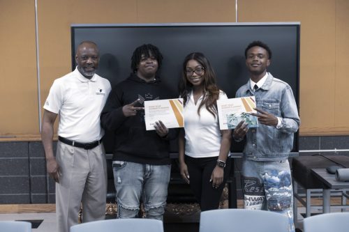 Four people pose indoors; two hold certificates and smile for a group photo in front of a screen.