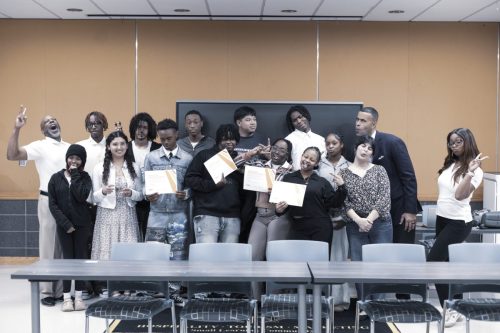 A group of students and adults pose playfully together, some holding certificates, in a classroom setting.