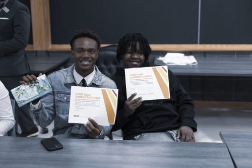 Two smiling students sit at a table holding up certificates and posing for the camera in a classroom.