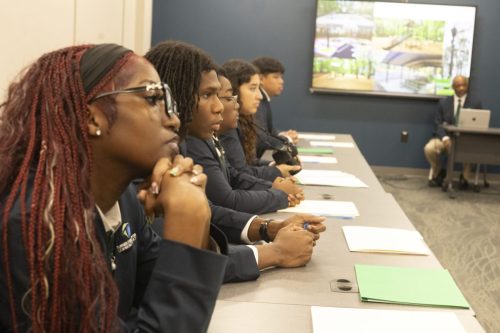 Students in business attire sit attentively at a conference table, listening to a presentation.
