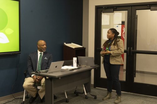 Two people in a meeting room; one is seated at a desk with laptops, the other is standing and speaking.